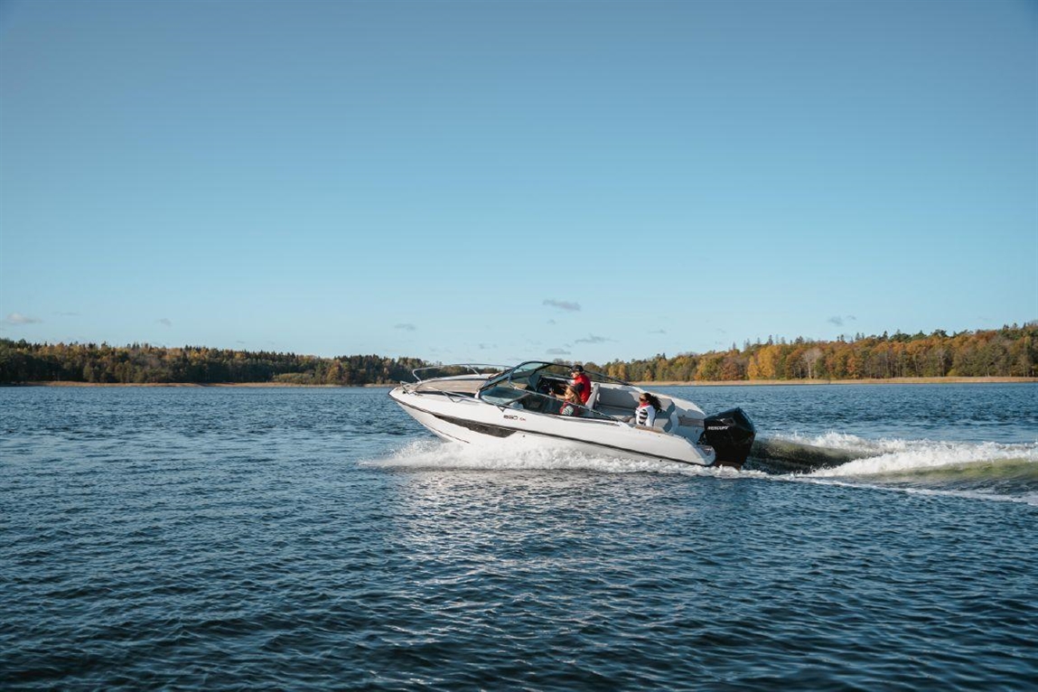 A white speedboat with four people is skimming across a calm blue lake on a sunny day with trees in the background.
